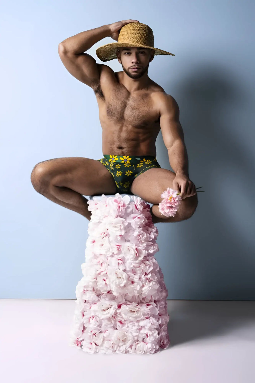 A man wearing a male power 145-285 petal power pouch short in green daisy print sits on a rose pedestal