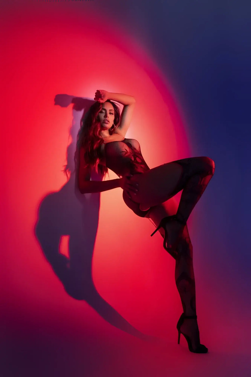 A woman poses in a black Mapale 1154 snake fishnet bodysuit with lace detailing and matching heels against a red blue backdrop