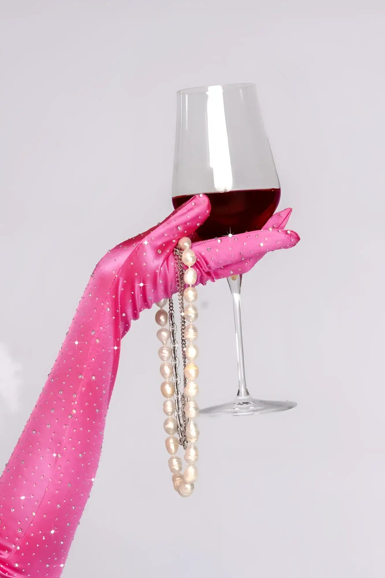 A hand in pink satin rhinestone opera glove holds a wine glass with red wine and pearl necklaces draped over it