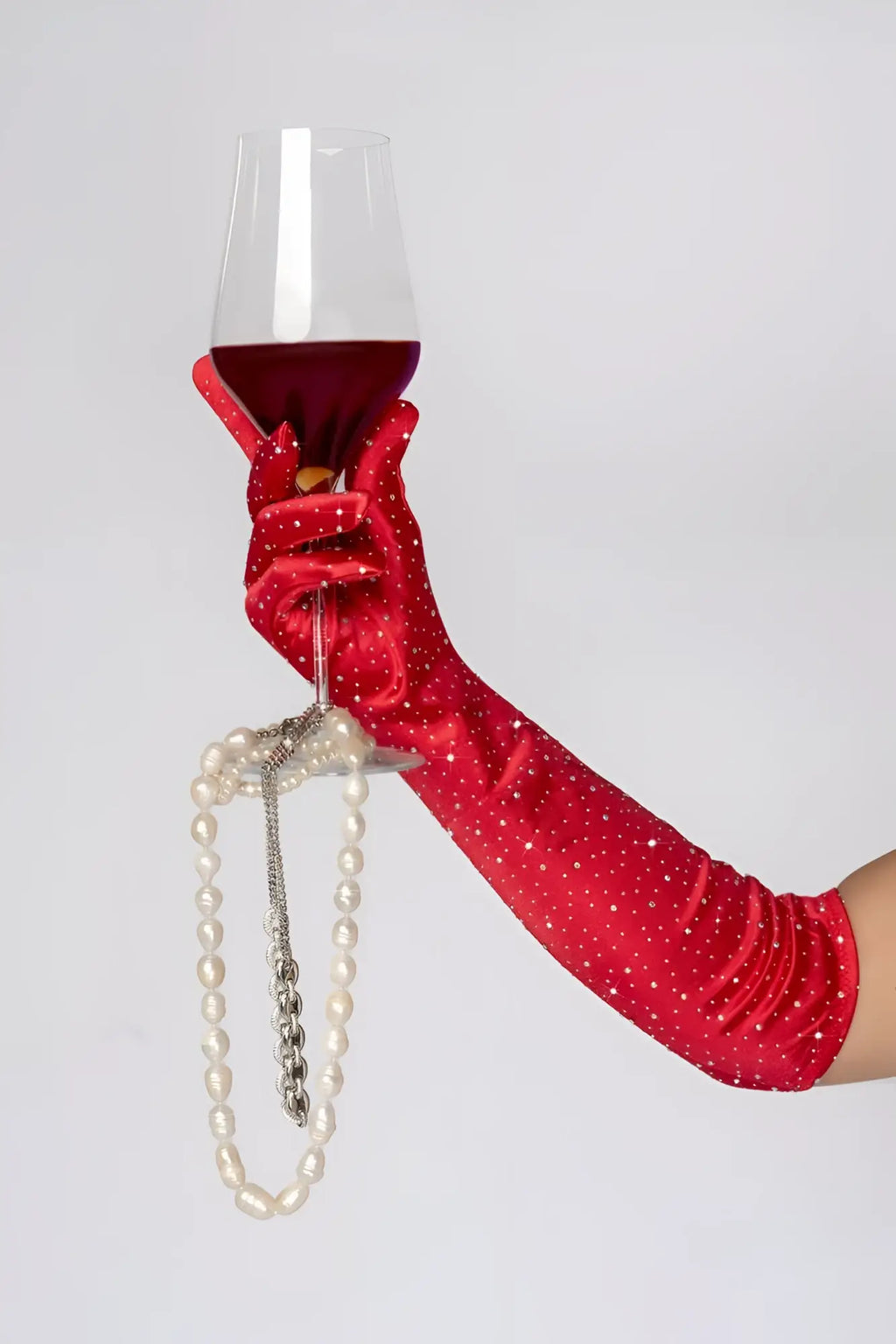 A hand wearing mapale 135 satin rhinestone opera gloves in red holds a wine glass with a pearl necklace on the stem