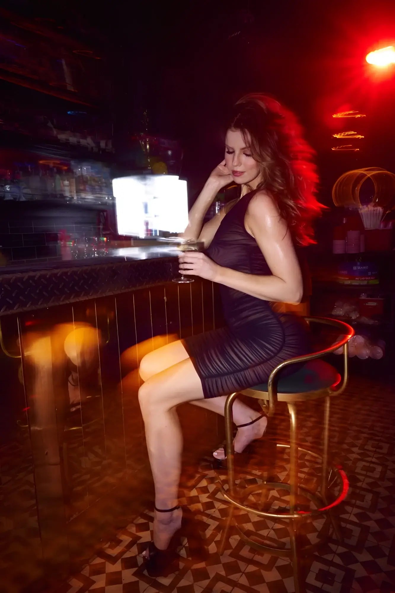 A woman in a form-fitting black mapale 40002 dress sits on a gold stool holding a glass of wine