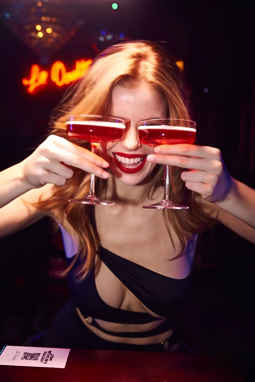 A woman with blonde hair holds two coupe glasses with red liquid wearing a black Mapale 40006 dress