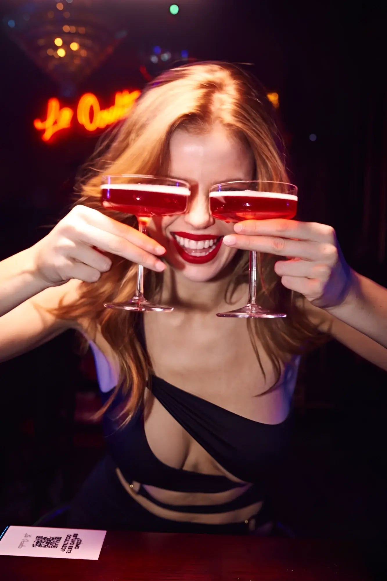 A woman with blonde hair holds two coupe glasses with red liquid wearing a black Mapale 40006 dress