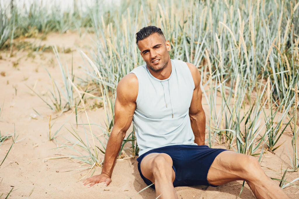 A man sits on sand wearing a light gray marcuse hero sleeveless hoodie tank top and navy blue shorts surrounded by grass