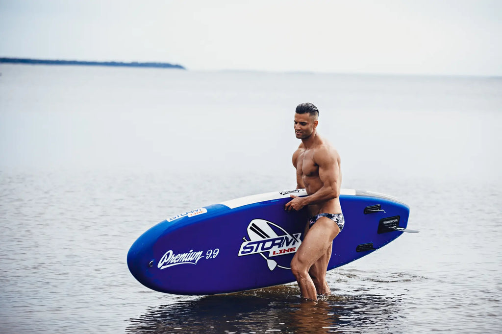 Blue inflatable surfboard with white branding and black traction pads featured with Marcuse Master bikini swimwear