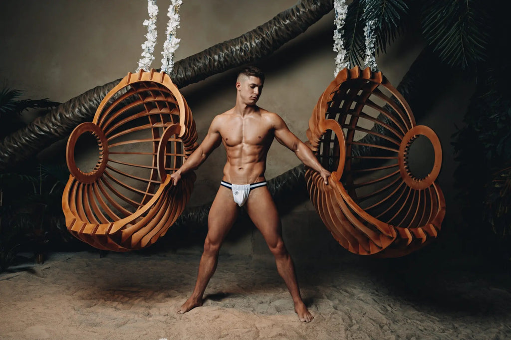 A man stands between two large wooden hanging swings wearing marcuse maximo jock underwear in warm brown