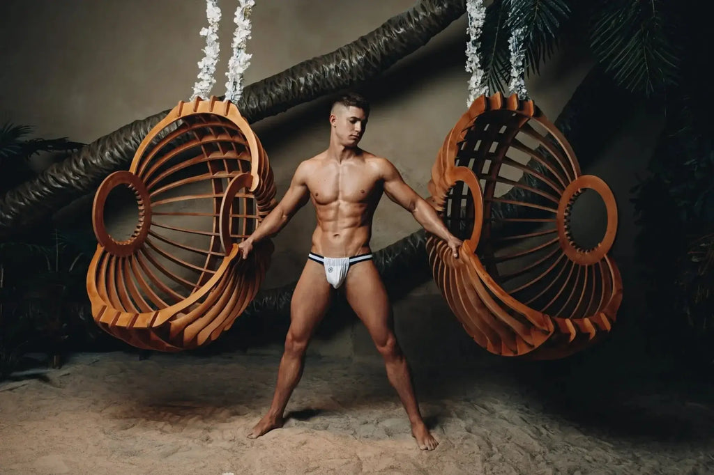 A man stands between two large wooden hanging swings wearing marcuse maximo jock underwear in warm brown
