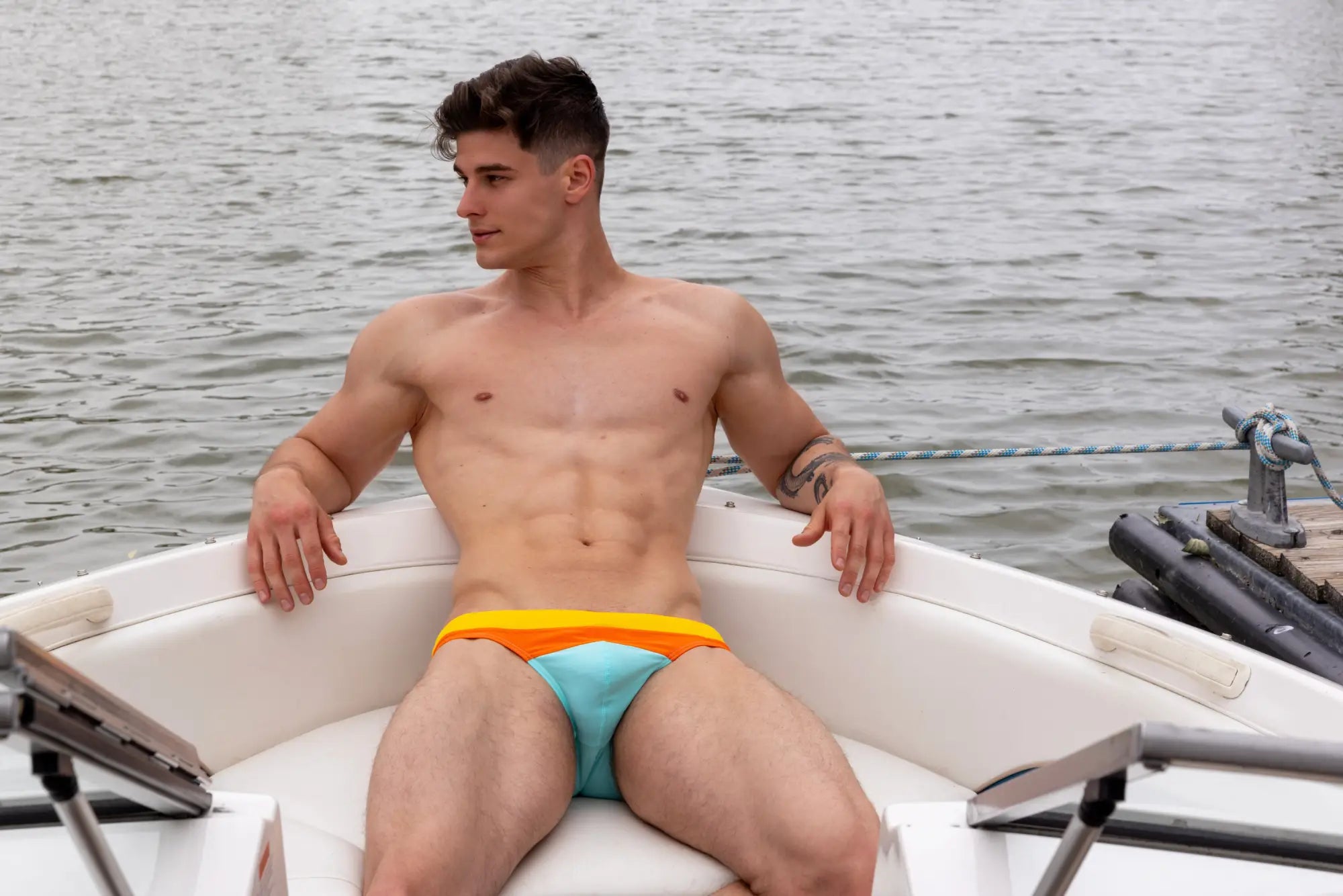 A man wearing Marcuse Seacoast bikini swimwear with vibrant two-tone swim trunks featuring an orange waistband and teal fabric