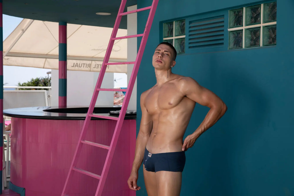 A man wearing navy blue Marcuse Waterboy boxer underwear stands against a teal wall with a pink ladder nearby