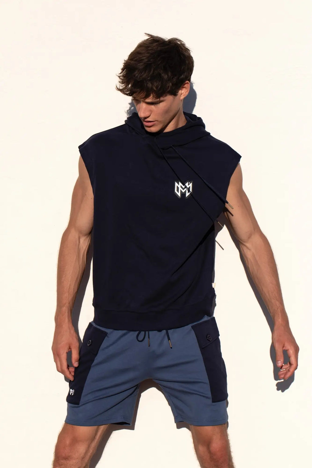Navy blue sleeveless hoodie with white logo on chest paired with marcuse zaiden cargo shorts for casual wear