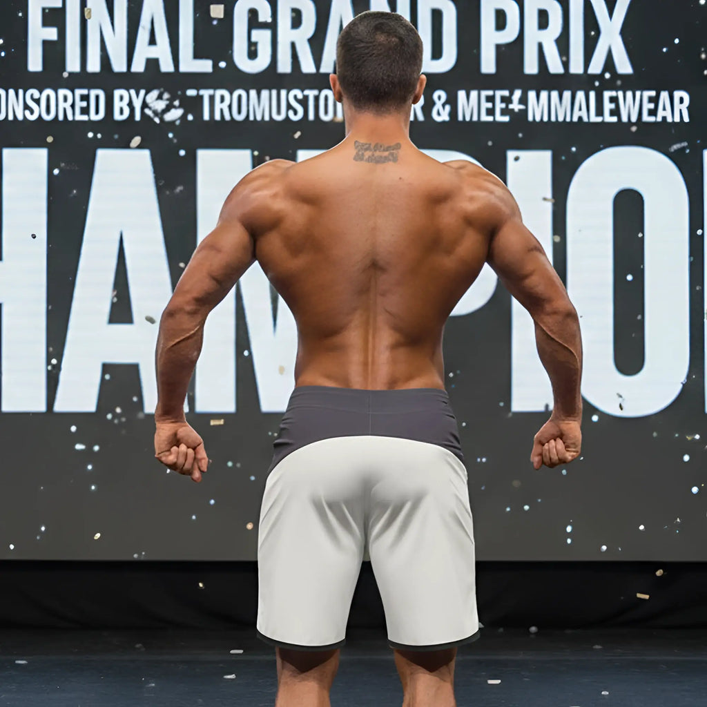 A muscular man stands on stage wearing gray MetroMaleWear MetroMuscleWear Cordoba physique board shorts with a dark waistband