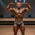 A muscular man with glossy tanned skin flexing arms in black metro musclwear elia muscle trunk competition attire