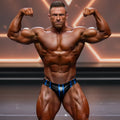 A muscular man flexing in black and blue striped metroMuscleWear stripe competition briefs on stage