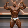 A muscular man flexing in a gold and white metro musclewear superior competition suit briefs on stage