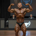A muscular man flexing his arms wearing black shiny MetroMaleWear MetroMuscleWear Zeus muscle trunk briefs on stage