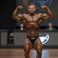A muscular man flexing his arms wearing black shiny MetroMaleWear MetroMuscleWear Zeus muscle trunk briefs on stage