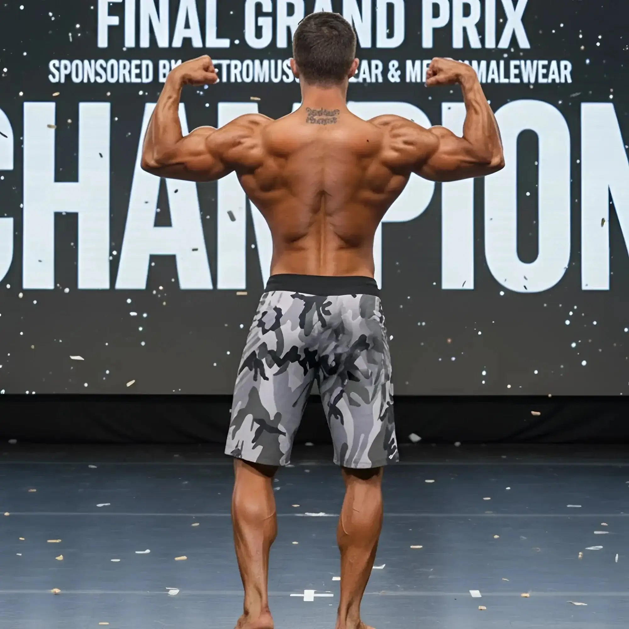 A muscular man wearing metroMalewear physique board short camo with black waistband stands on stage facing front