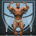 A muscular man wearing MetroMaleWear PosingWear hologram gold shiny competition briefs with number 38 on hip