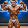 A muscular man wearing MetroMaleWear posingwear snake skin white and gray patterned competition briefs with number 18