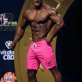 Bright pink MetroMaleWear physique board shorts with white drawstring and black shield logo on lower left leg