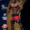 A muscular man wearing metro male wear physique board short red 4706-12 with a white bull logo on left leg