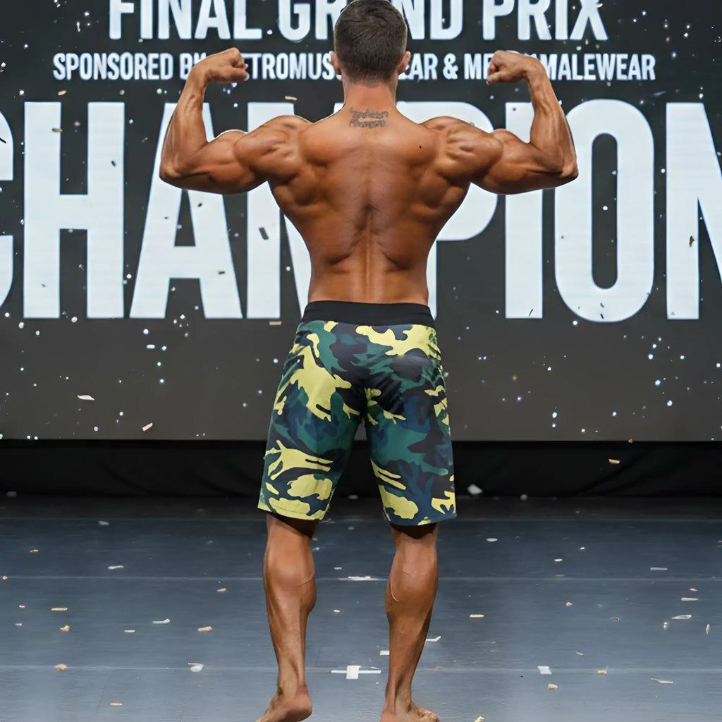 A muscular man stands on stage wearing metroMalewear physique board short yellow camo with green and yellow patterns