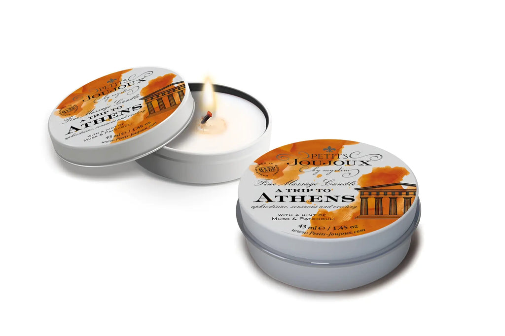 A lit white scented massage candle in a silver tin labeled petits joujoux a trip to athens with orange and black design