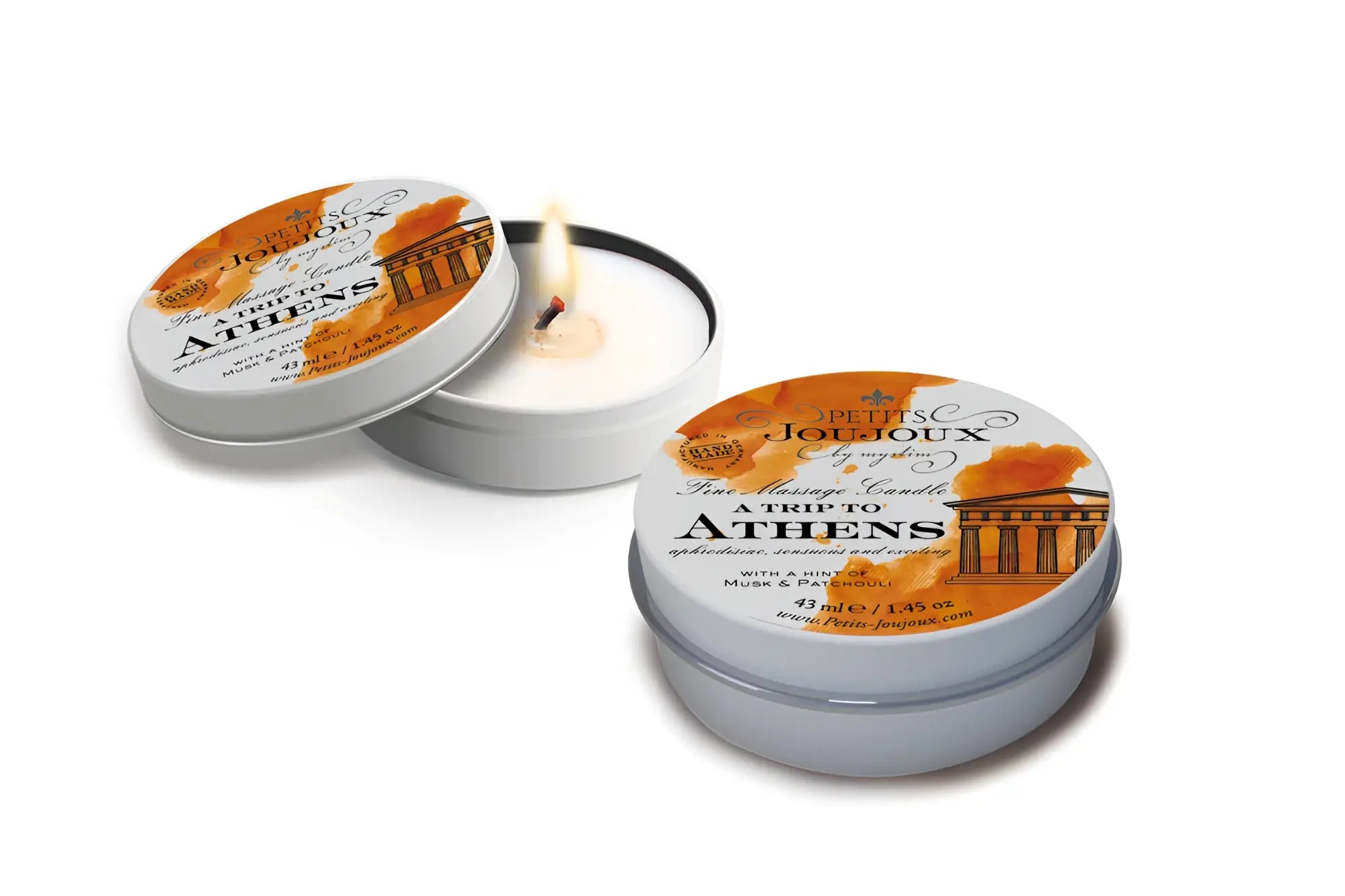 A lit white scented massage candle in a silver tin labeled petits joujoux a trip to athens with orange and black design
