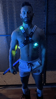 The man is wearing a black and blue rave cross harness with led-lit glowing green lights