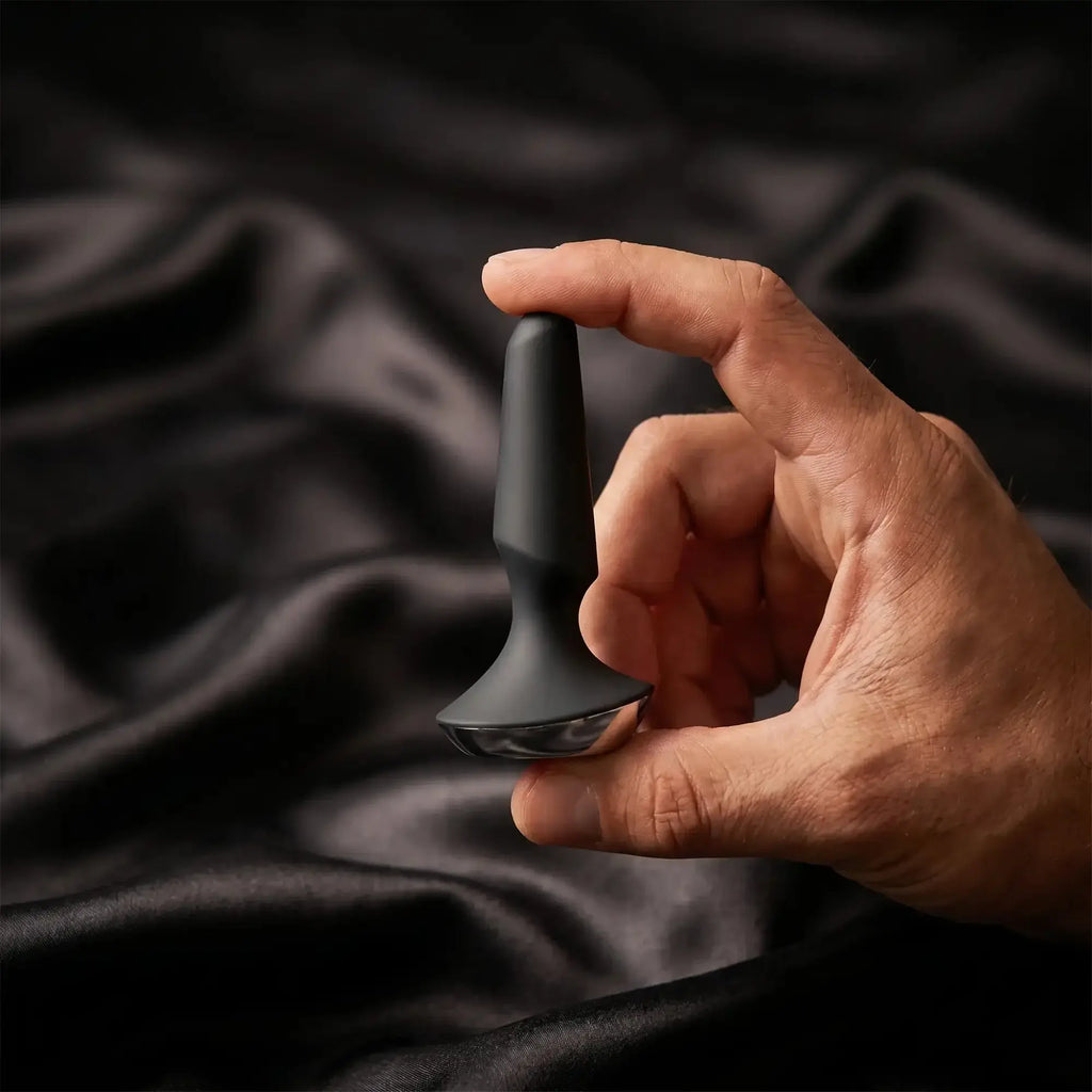 A man's hand holds a matte black silicone anal plug against soft dark satin — lifestyle image for the Anal collection