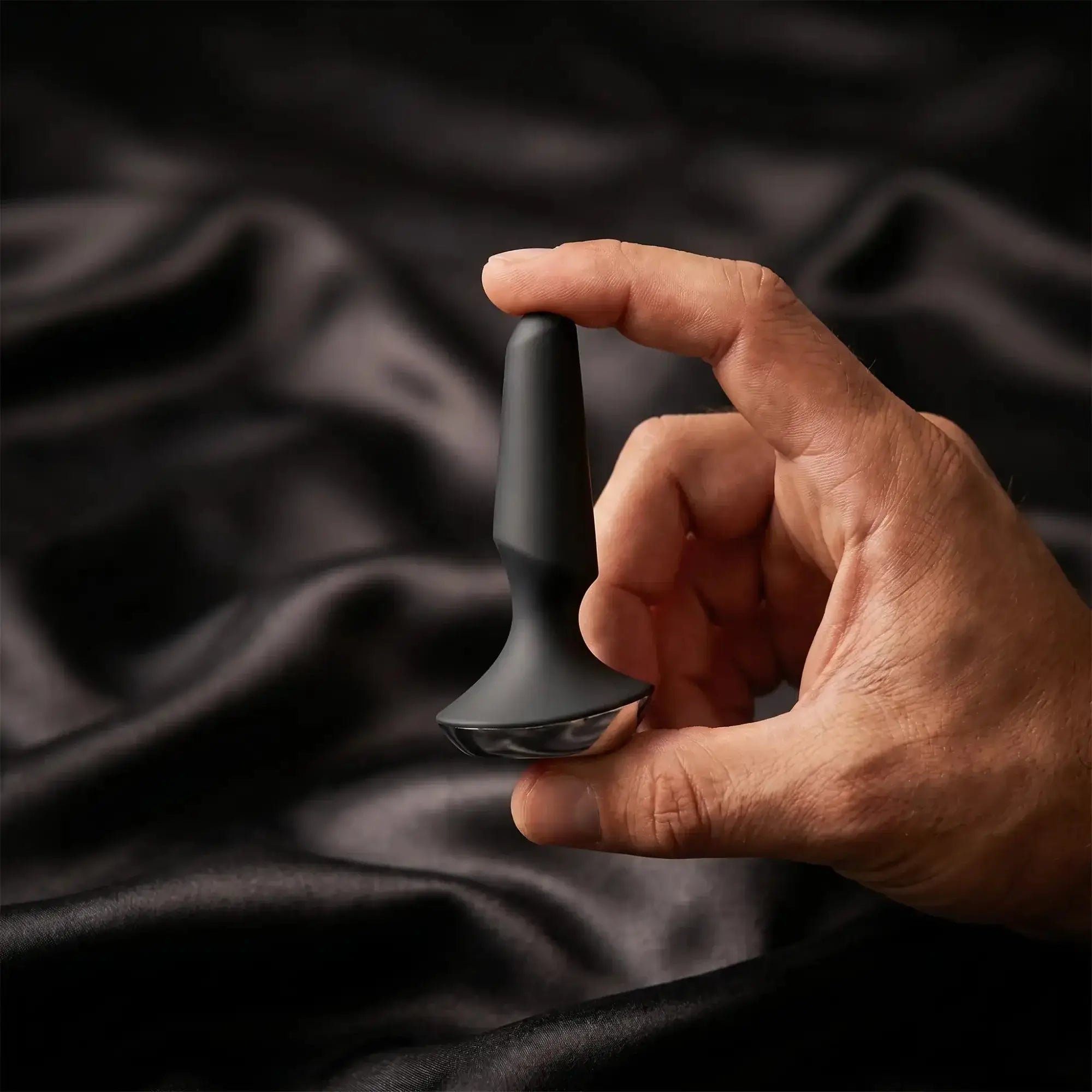 A man's hand holds a matte black silicone anal plug against soft dark satin — lifestyle image for the Anal collection