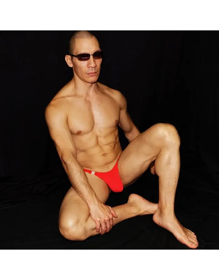 Red thong underwear by Arroyman featuring a snap closure worn by a model in a seated pose against a dark background.