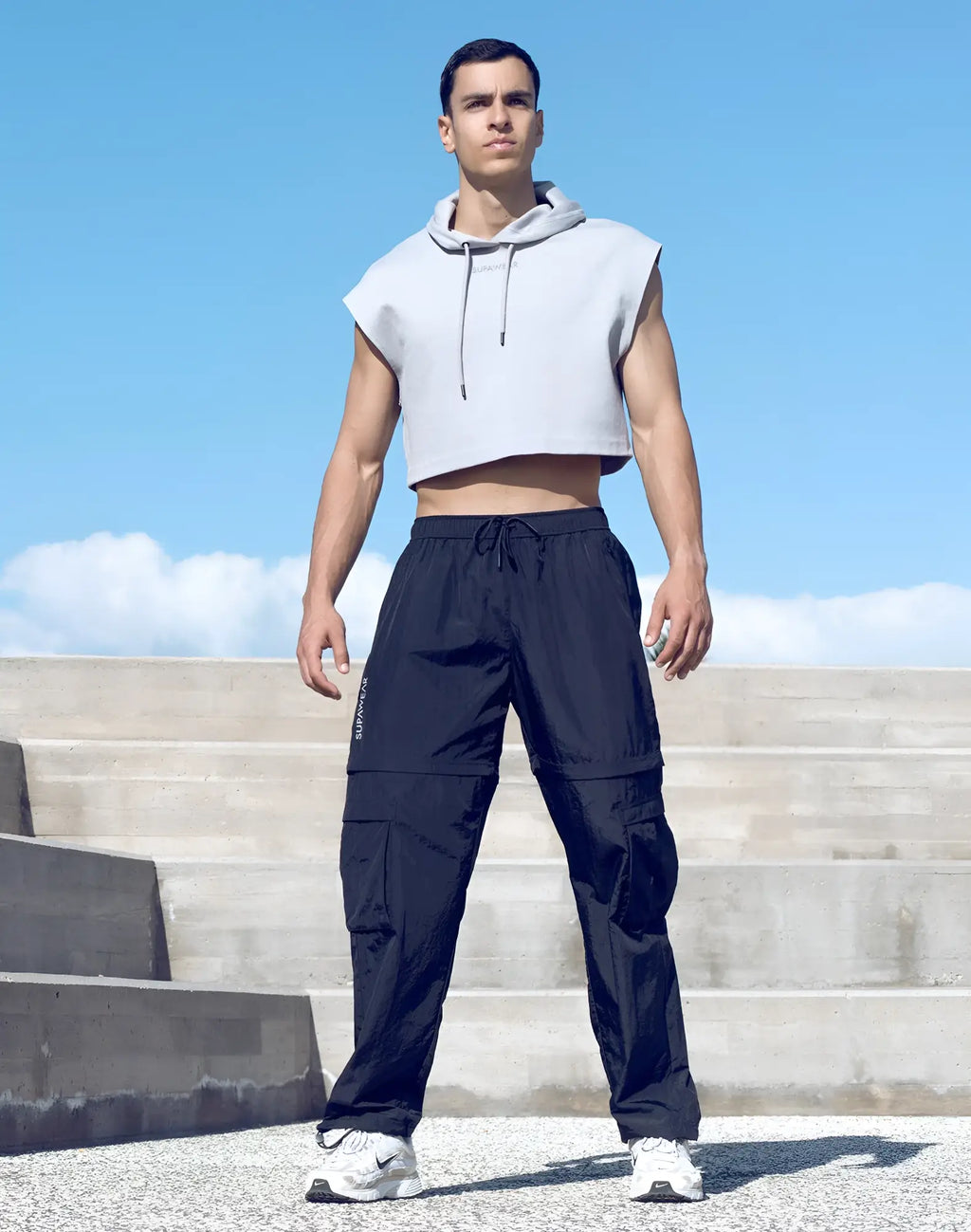 A man wearing a supawear grey sleeveless cropped hoodie with drawstrings and black cargo pants with pockets