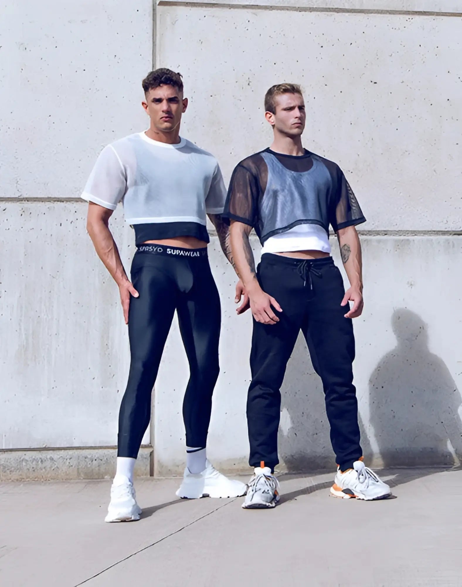 Two men model supawear mesh oversized cropped tees in white with black leggings and dark joggers for athletic wear