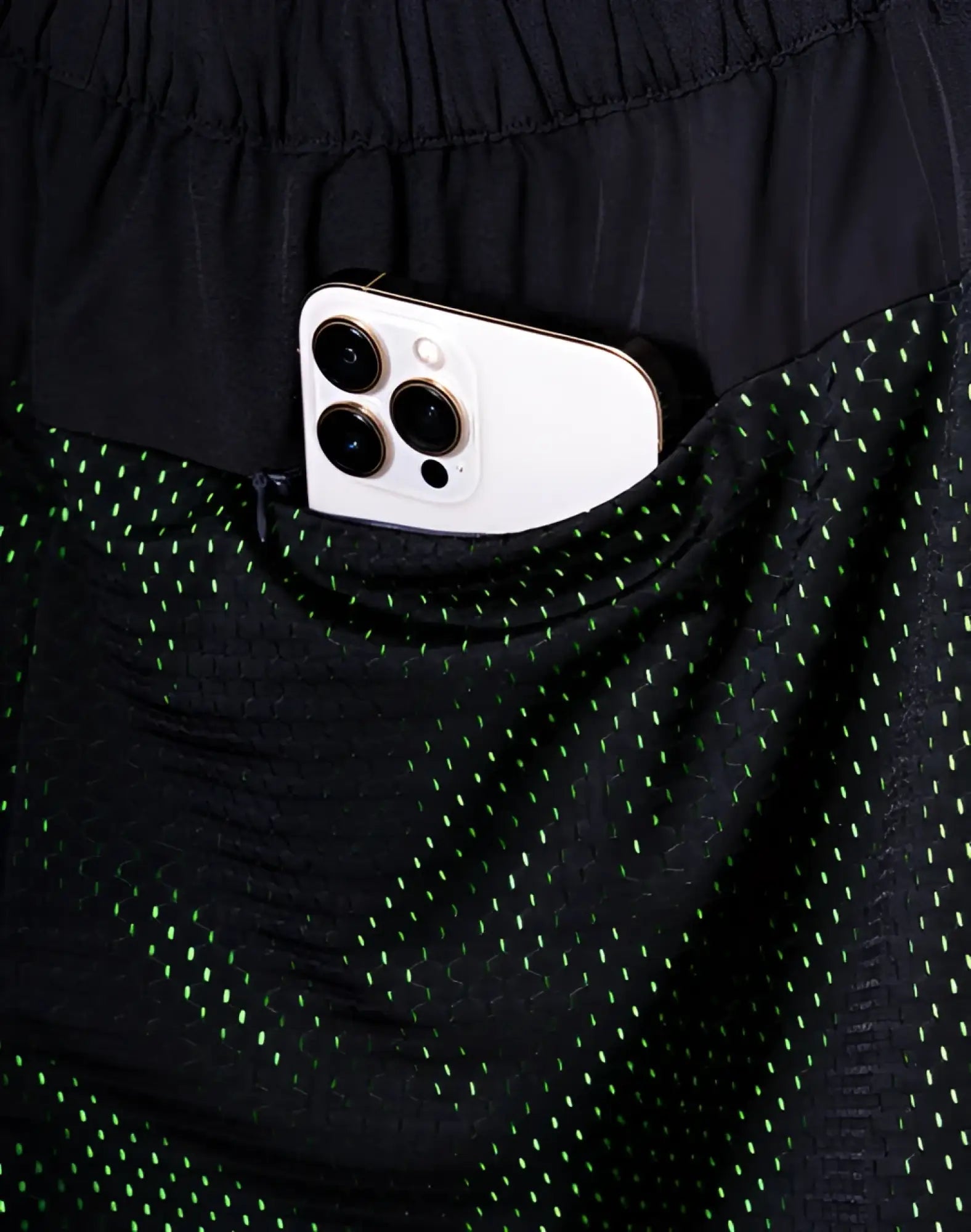 A white iphone 13 pro with triple-lens camera peeking from the side pocket of black supawear neon mesh shorts