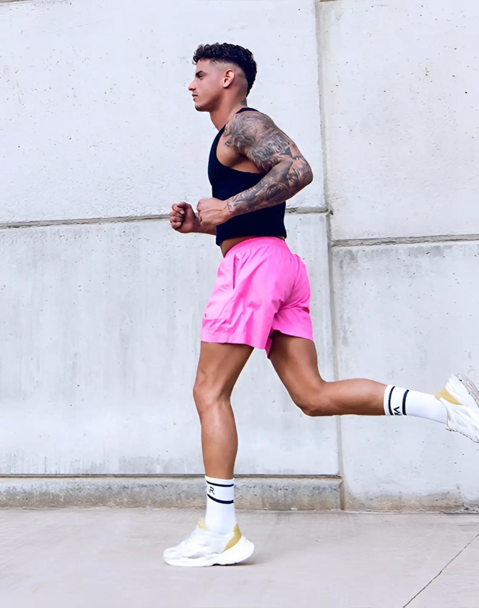 A man wearing supawear ribbed cropped singlet black with pink shorts and white sneakers walks by a concrete wall