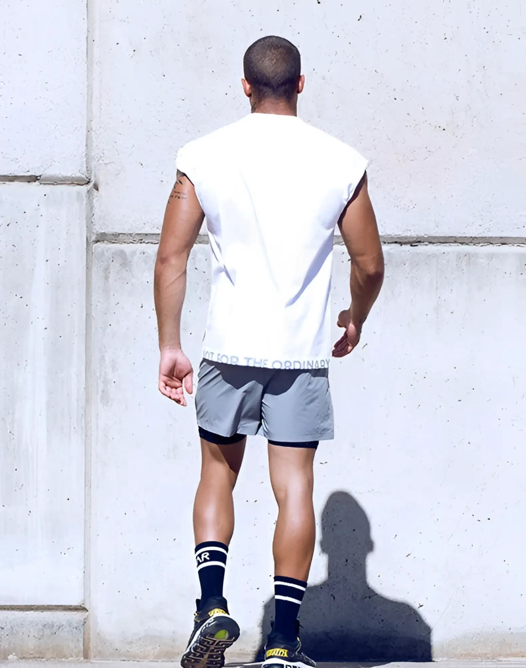 A man wearing a white SUPAWEAR wide-cut tank with for the ordinary waistband, gray shorts, and black striped socks