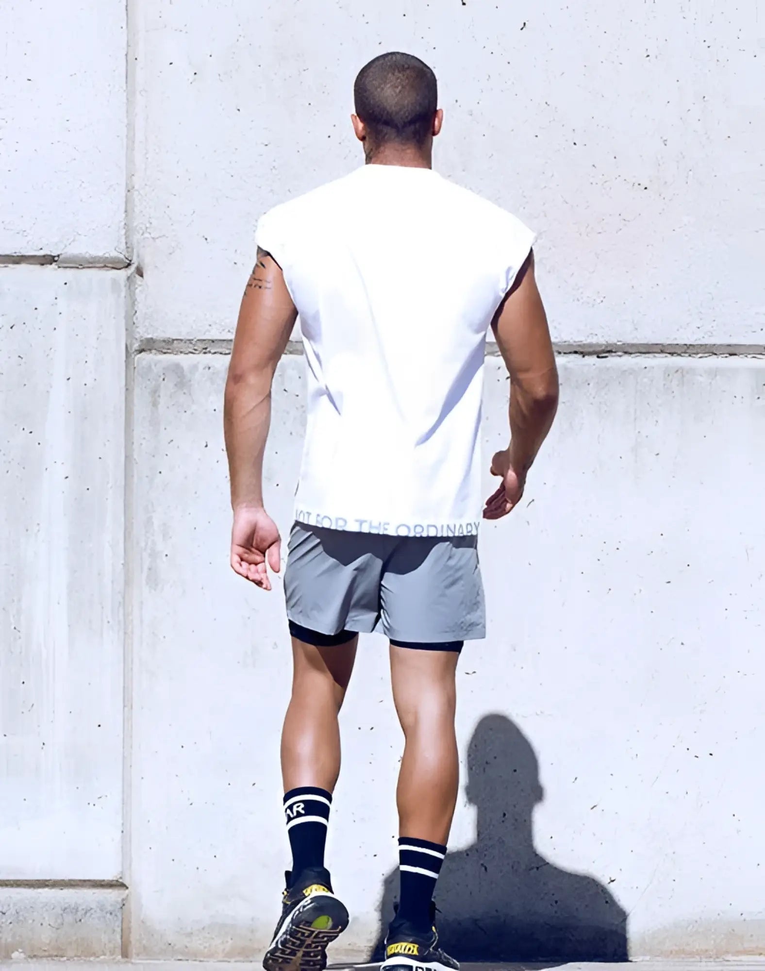 A man wearing a white SUPAWEAR wide-cut tank with for the ordinary waistband, gray shorts, and black striped socks