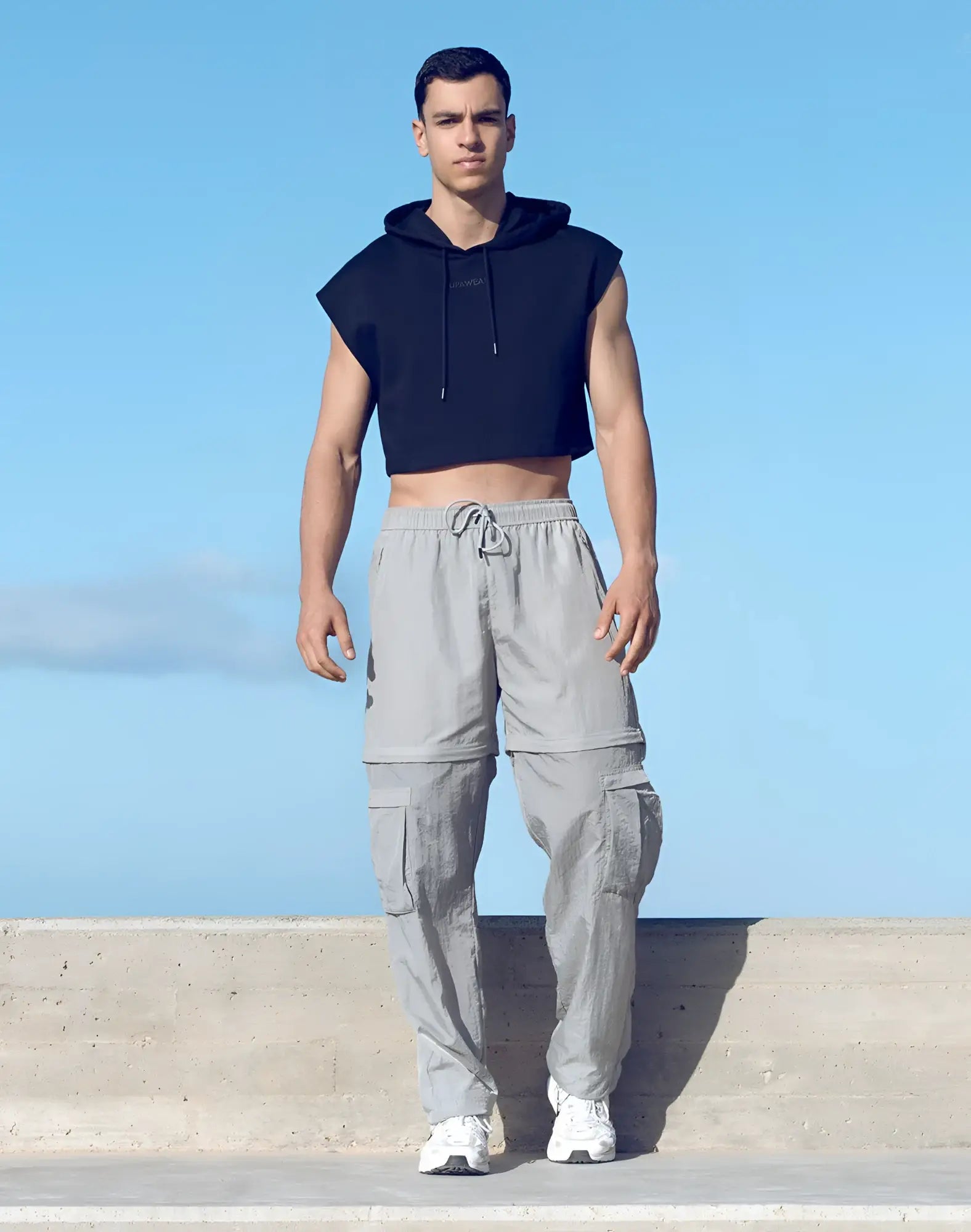 A man wearing a black sleeveless cropped hoodie and grey supawear zip-off trouser cargo pants with multiple pockets