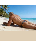 Model wearing Arroyman grey print beach thong swimwear lying on the sand by the ocean showcasing the back design.