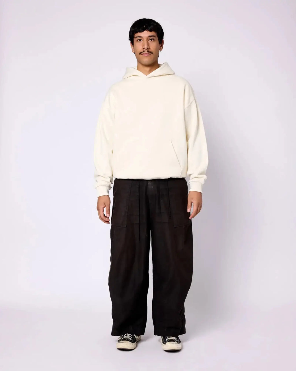 A man wears a plain, off-white, oversized cotton hoodie with a front kangaroo pocket and rib-knit cuffs.