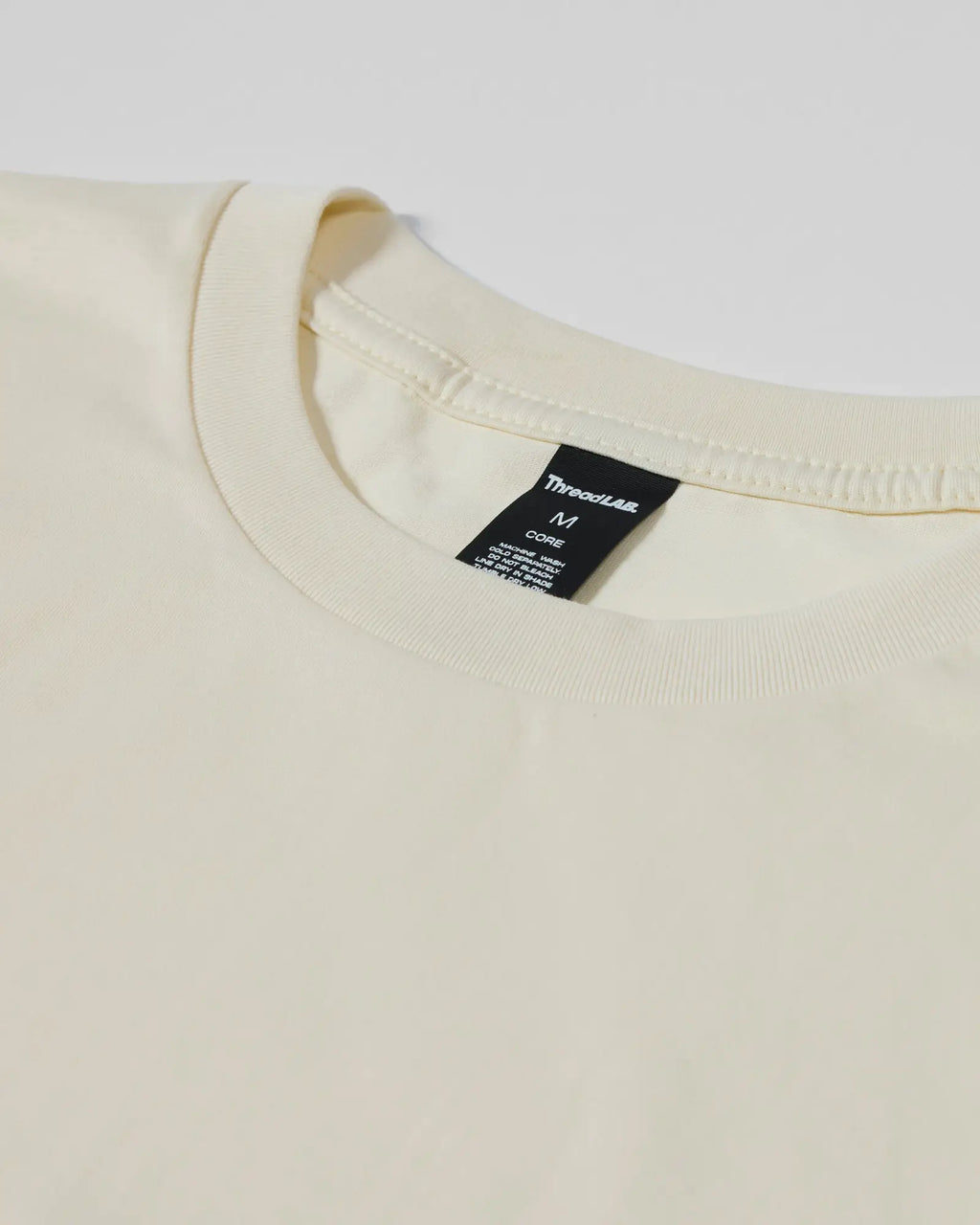 Close-up of a cream-colored crew neck T-shirt with a black ThreadLab brand tag at the neckline.