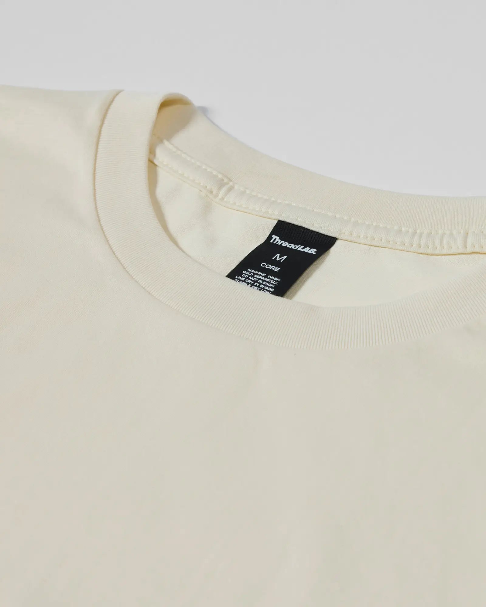 Close-up of a cream-colored crew neck T-shirt with a black ThreadLab brand tag at the neckline.