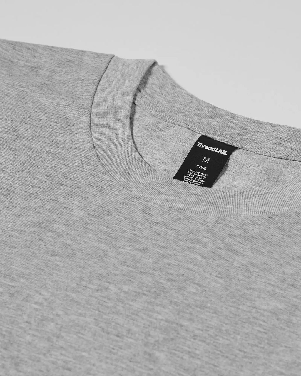 Close-up of a heather grey crew neck Core Tee with a black size M neck label by ThreadLAB.