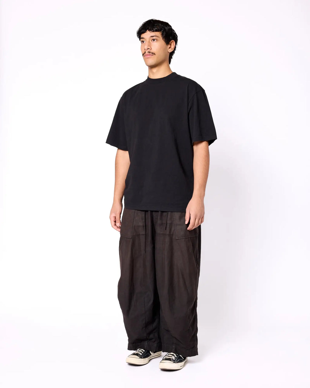 A man wearing a black, relaxed-fit crewneck t-shirt with short sleeves, styled with dark, wide-leg trousers.