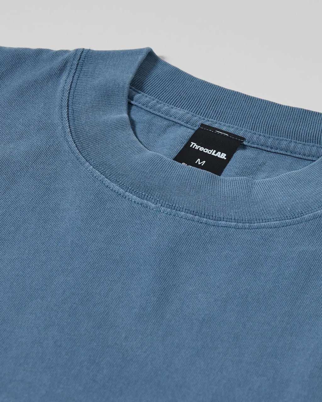 Close-up of a dusty blue crewneck t-shirt with a rib-knit collar and ThreadLAB brand tag in size medium.