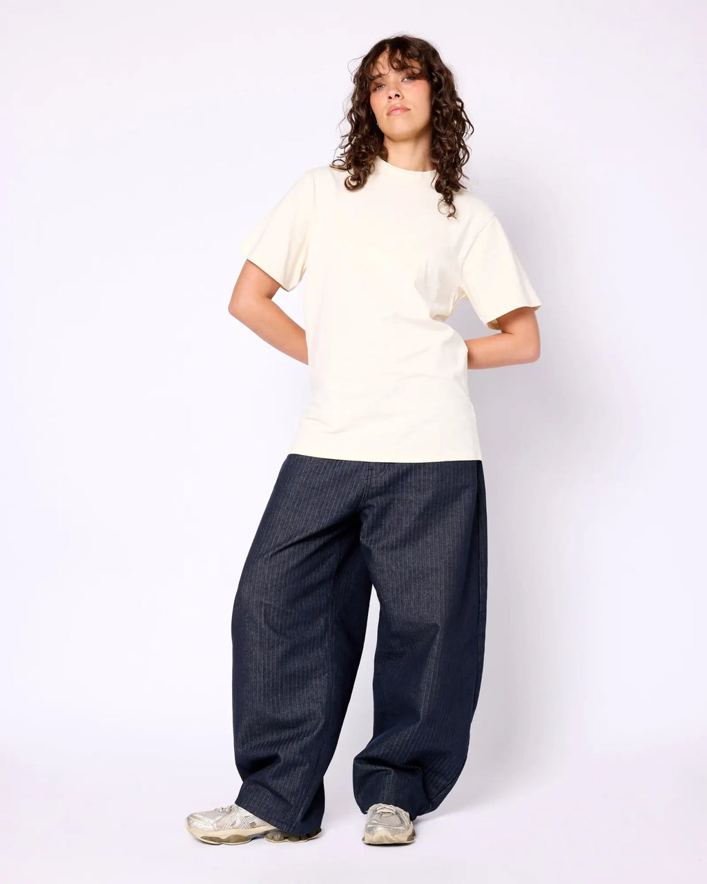 A model wears a cream-colored, crew-neck short-sleeve tee with loose-fit dark pinstripe trousers and silver sneakers.