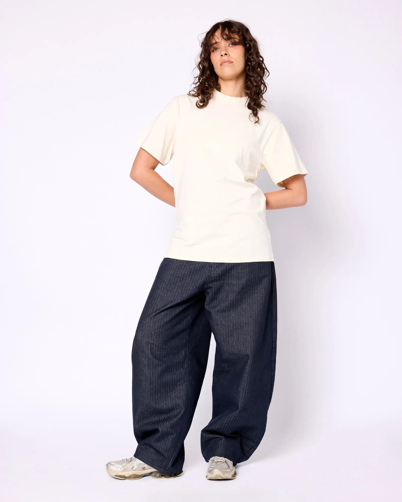 A model wears a cream-colored, crew-neck short-sleeve tee with loose-fit dark pinstripe trousers and silver sneakers.