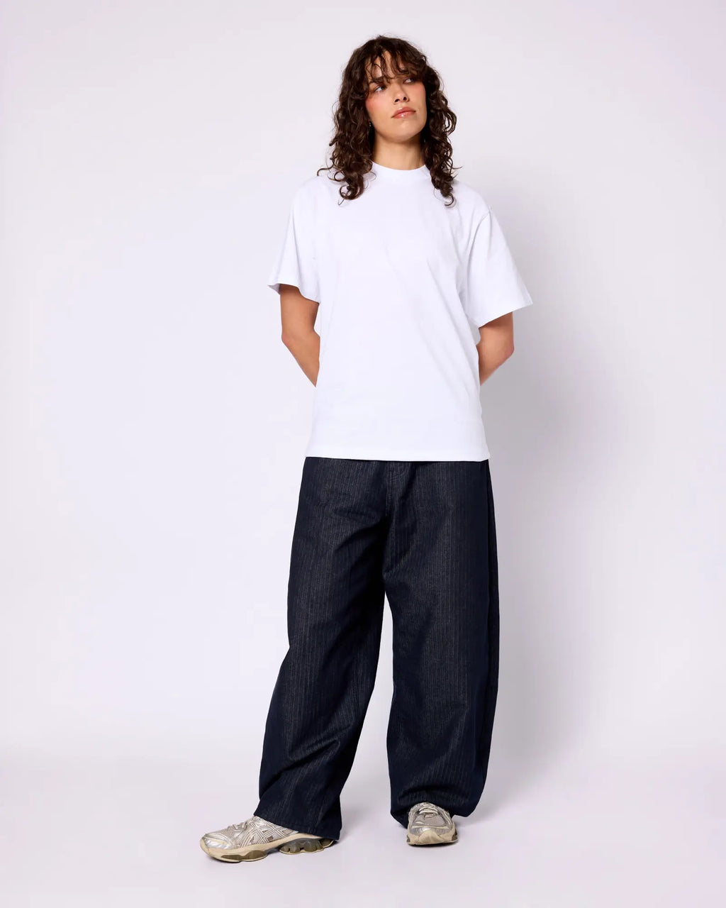 Model wears a clean white crewneck tee with a boxy, relaxed fit, paired with dark, wide-leg pinstripe denim pants.