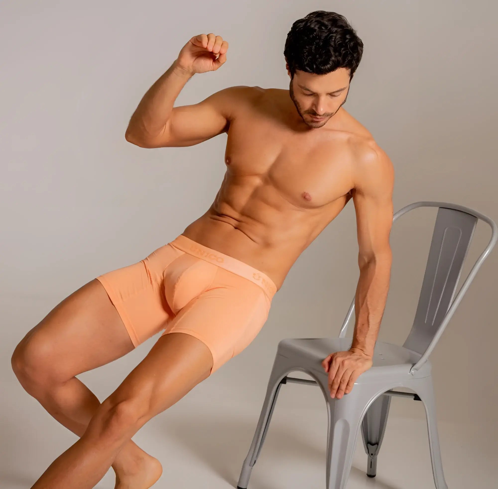 Lifestyle shot of a man wearing Unico Classic Boxer Briefs in Apricot, posing with a gray chair.